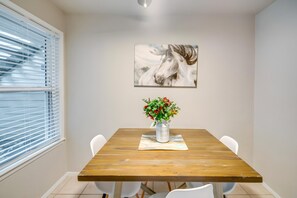 Uma sala de jantar com uma mesa de madeira, cadeiras brancas, um vaso de flores e um quadro de cavalos na parede.