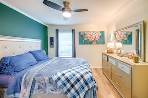 Une chambre à coucher comprenant un lit, une commode, un ventilateur de plafond et deux tableaux à motifs floraux accrochés au mur.