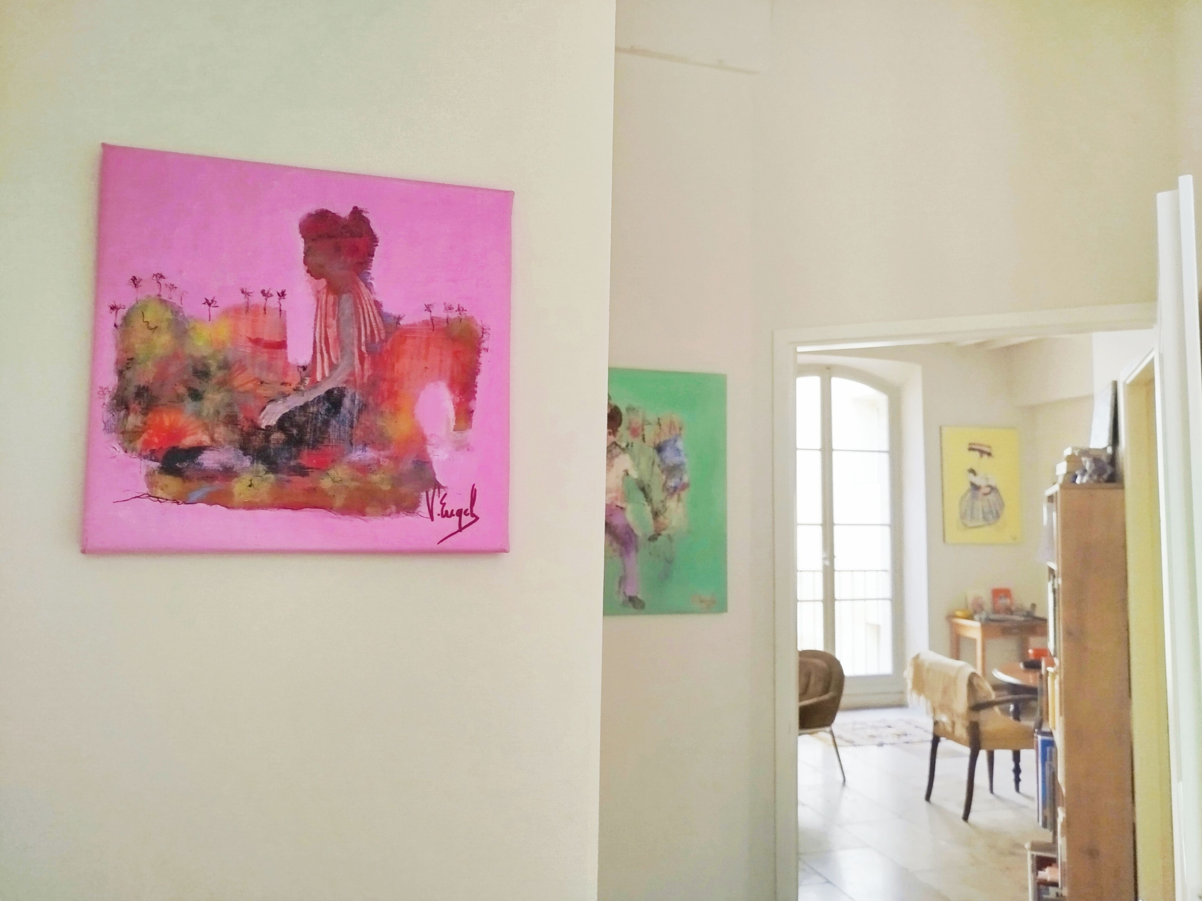 A room with a pink abstract painting on the wall, a green abstract painting, and a view of a dining area through an open doorway.