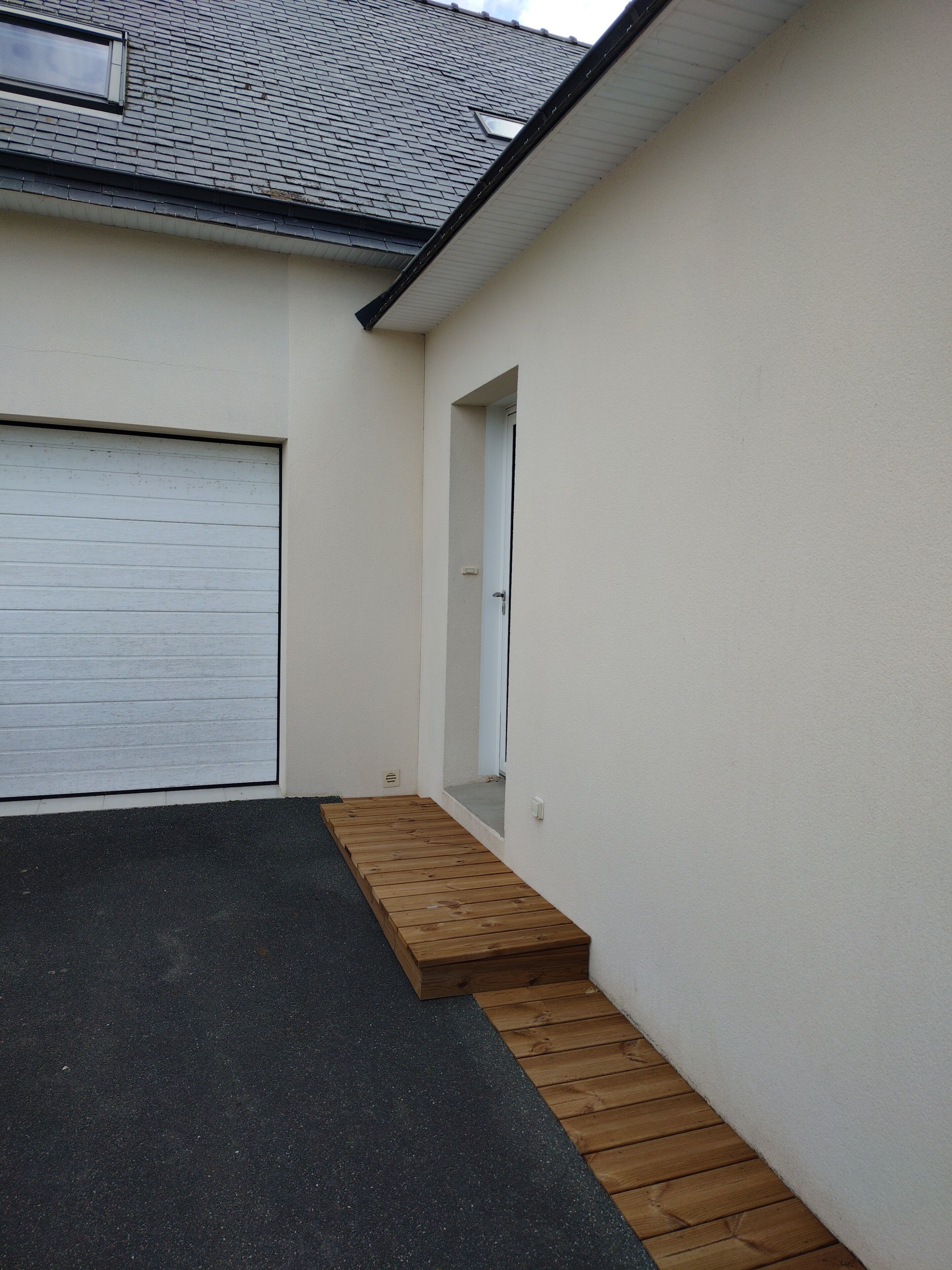 A garage with a white door, a wooden ramp leading to a driveway, and a small step leading to a door.