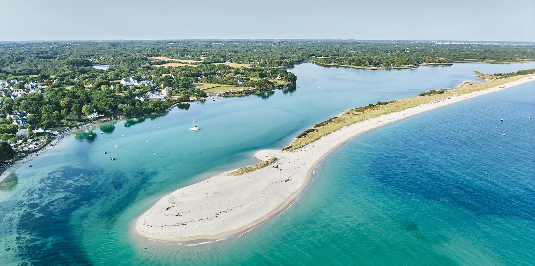 An aerial view of a coastal area with a sandy beach, clear turquoise waters, and a residential area with houses and greenery in the background.
