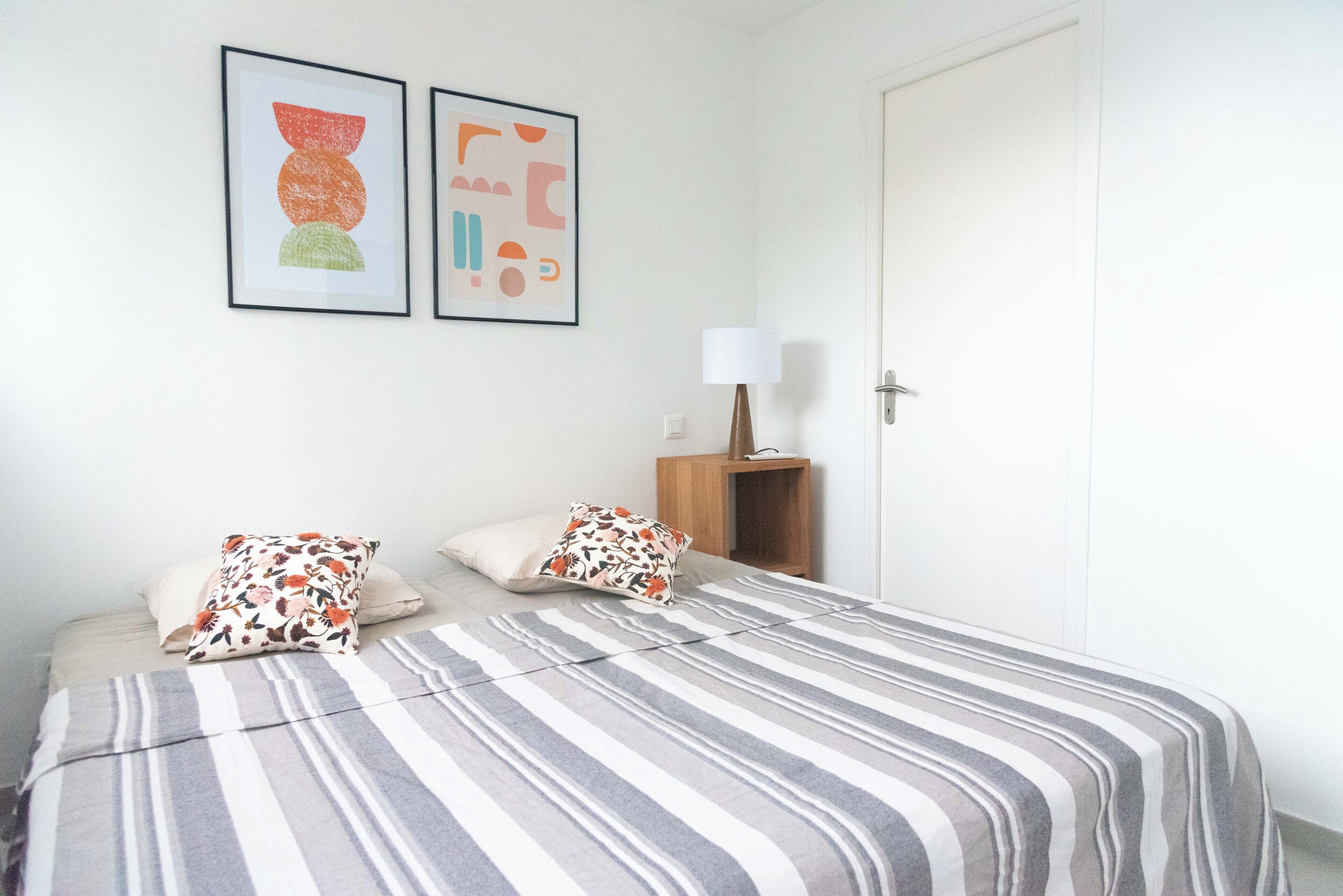 Ein Schlafzimmer mit einem Bett, Kissen, einem Nachttisch, einer Lampe und zwei gerahmten Kunstwerken an der Wand.