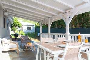 Eine überdachte Terrasse mit weißen Möbeln, einem Esstisch und Blick auf ein Haus und Grünflächen.