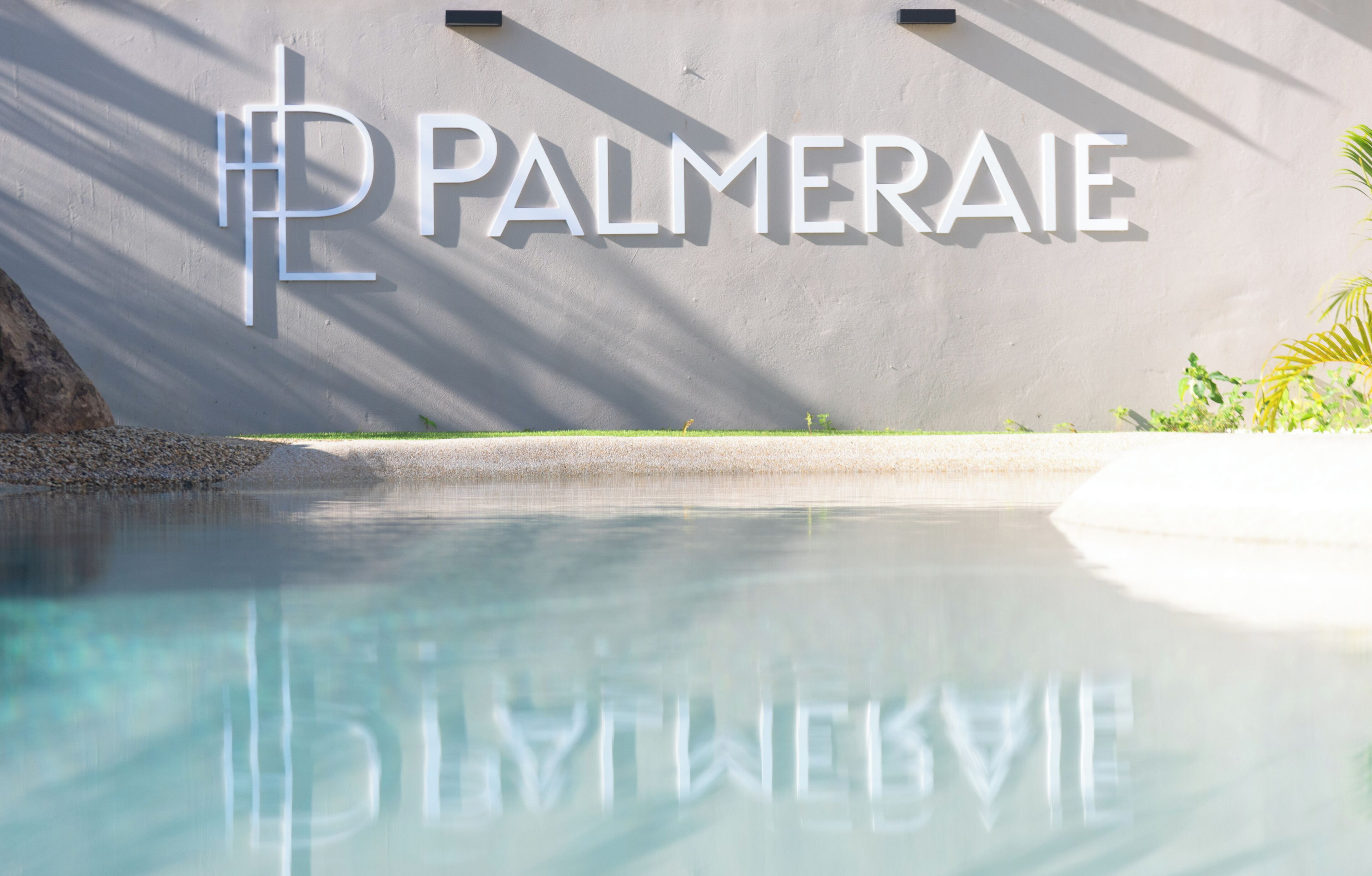 Ein klares Schwimmbecken, in dessen Spiegelung der Schriftzug 'PALMERAEI' an der Wand zu sehen ist.