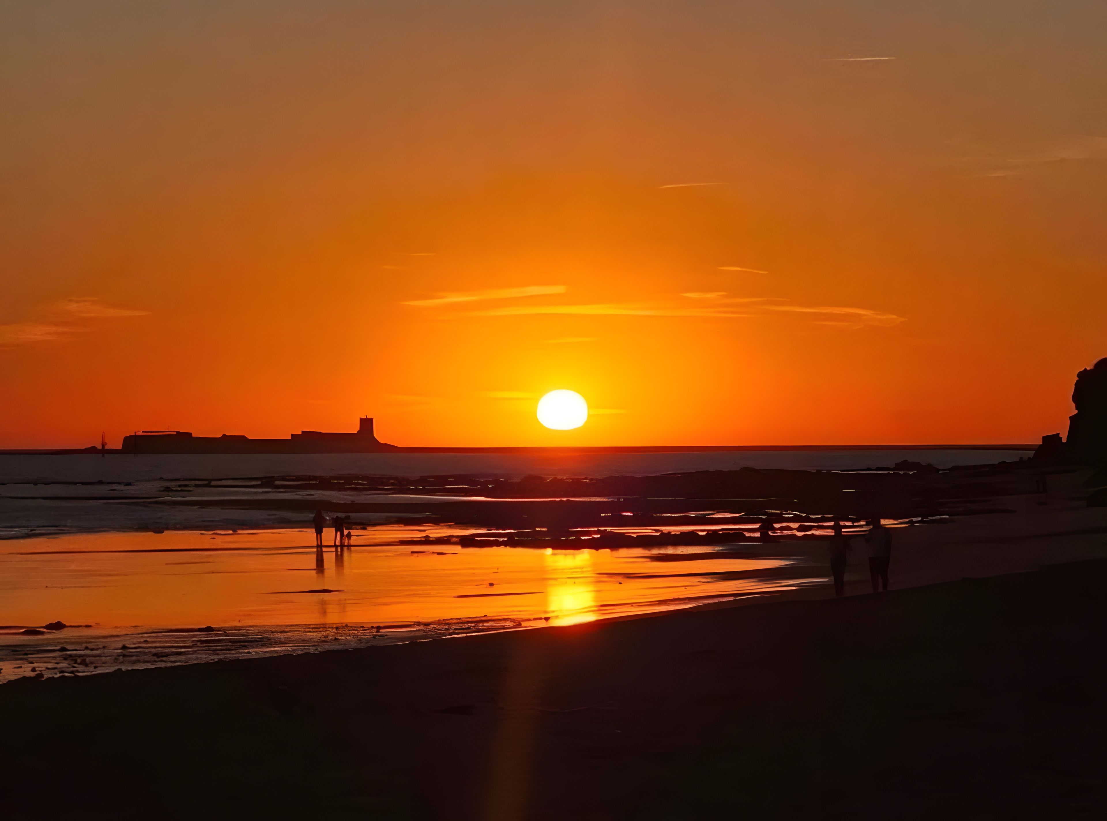 Un coucher de soleil sur une plage, avec un ciel dégagé, une eau calme et des bâtiments visibles au loin.
