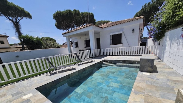 Uma piscina com um deck ladrilhado, cerca branca e um edifício com telhado ladrilhado ao fundo.