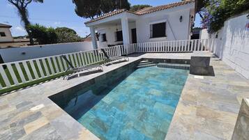 Una piscina con bordo piastrellato, una recinzione bianca e un edificio con tetto in tegole sullo sfondo.