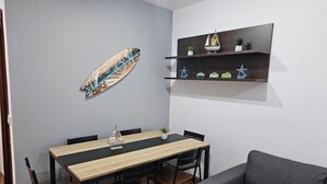 Uma área de refeições com mesa e cadeiras de madeira, um sofá cinza, uma prateleira fixada na parede com objetos decorativos e uma obra de arte com tema de prancha de surf.