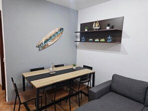 Een eethoek met een houten tafel en stoelen, een grijze bank, een wandplank met decoratieve objecten, en een surfboard kunstwerk.