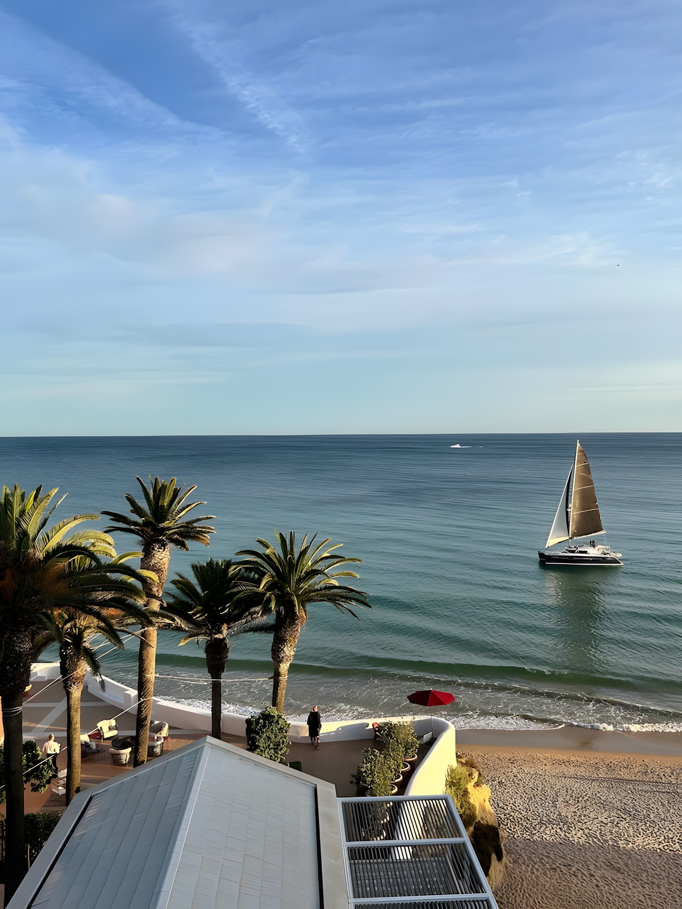Una platja amb palmeres, un veler i un edifici amb teulada inclinada.