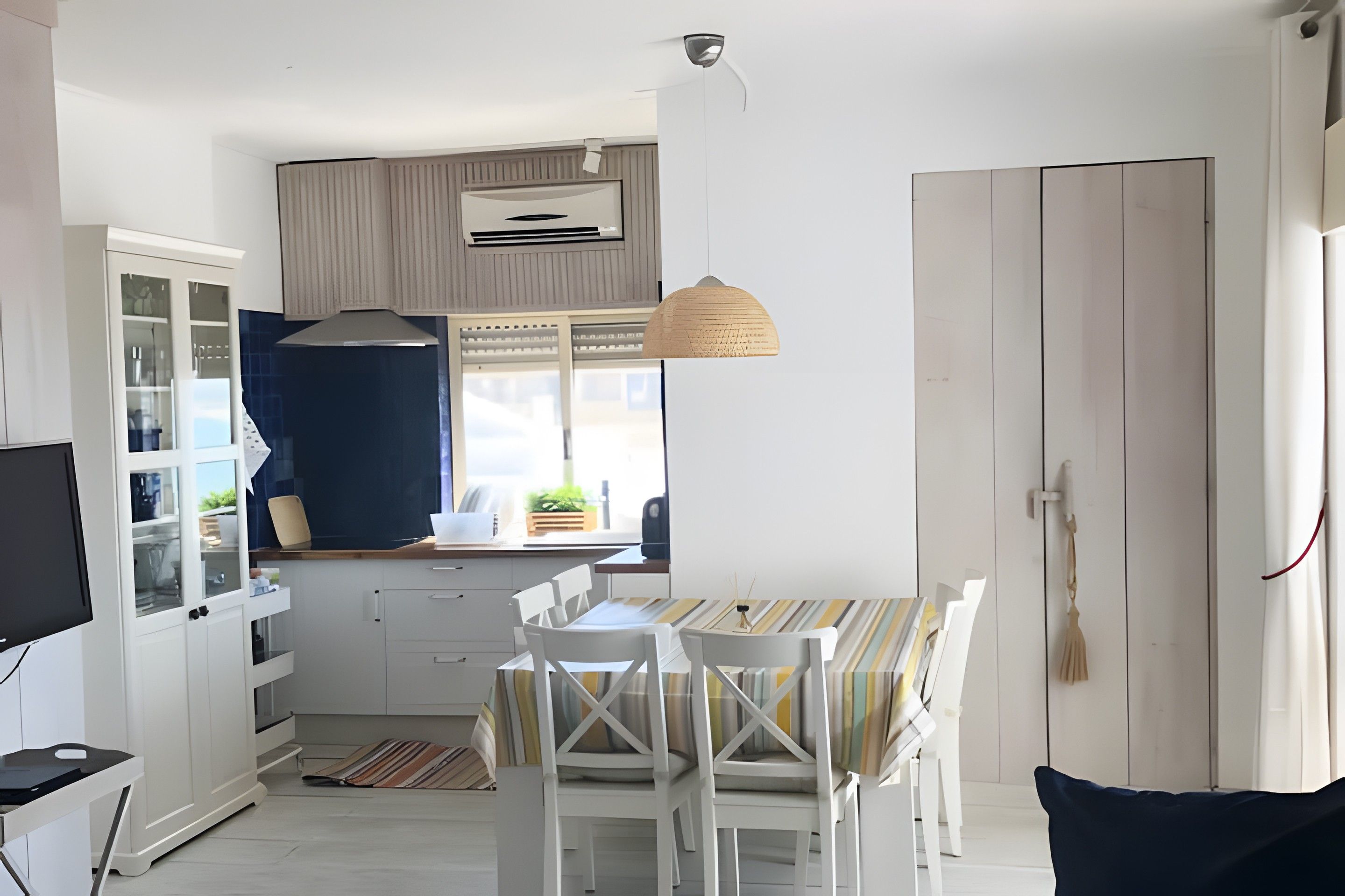 Cuina compacta amb armaris blancs, una taula de dinar amb mantell rayat i una làmpada penjant.