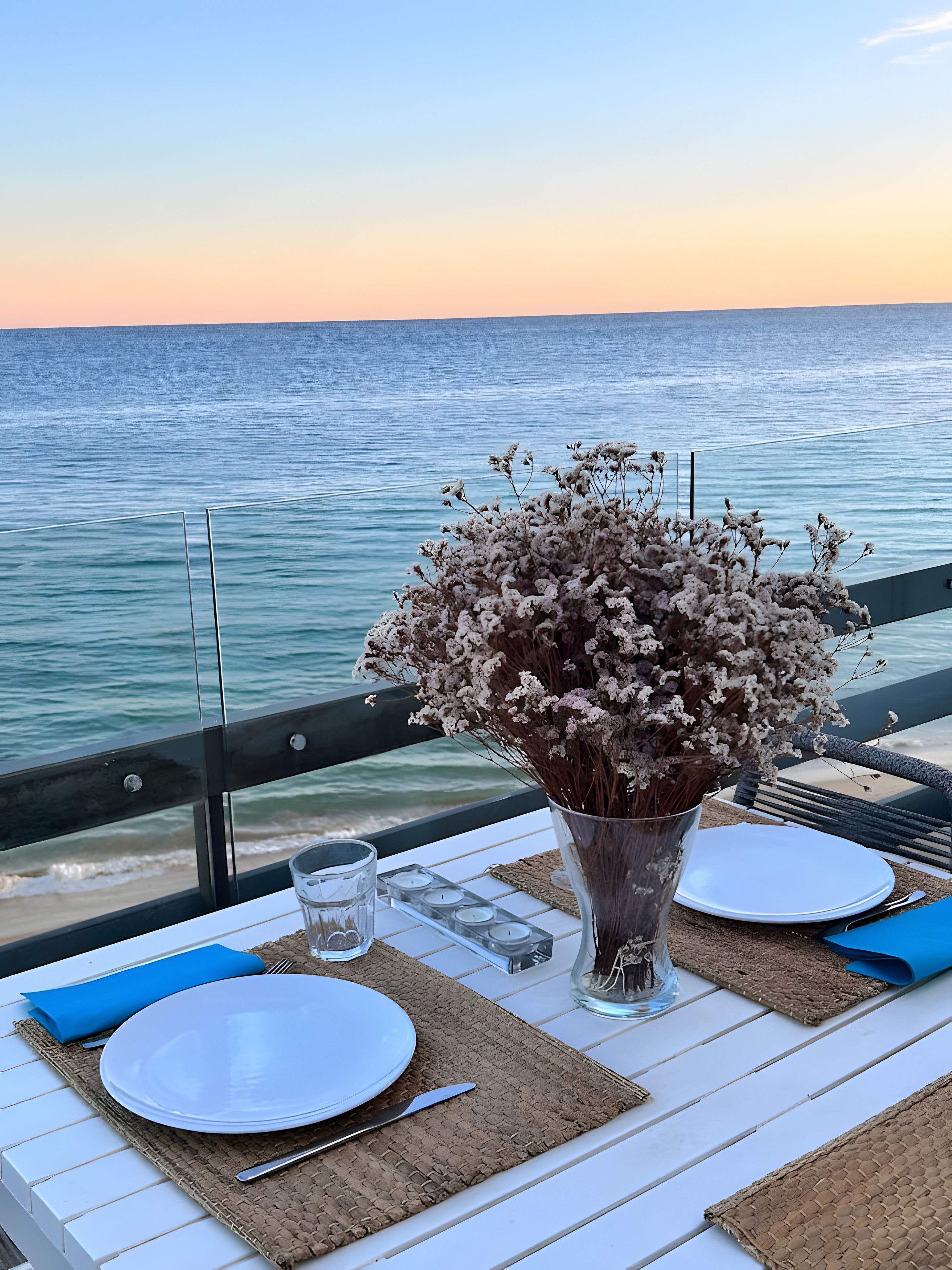 Taula posta amb plats, coberts i espelmes, amb vista a un mar tranquil.