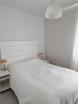 Quarto com cama branca, mesa de cabeceira branca, luminária e luz de teto branca.