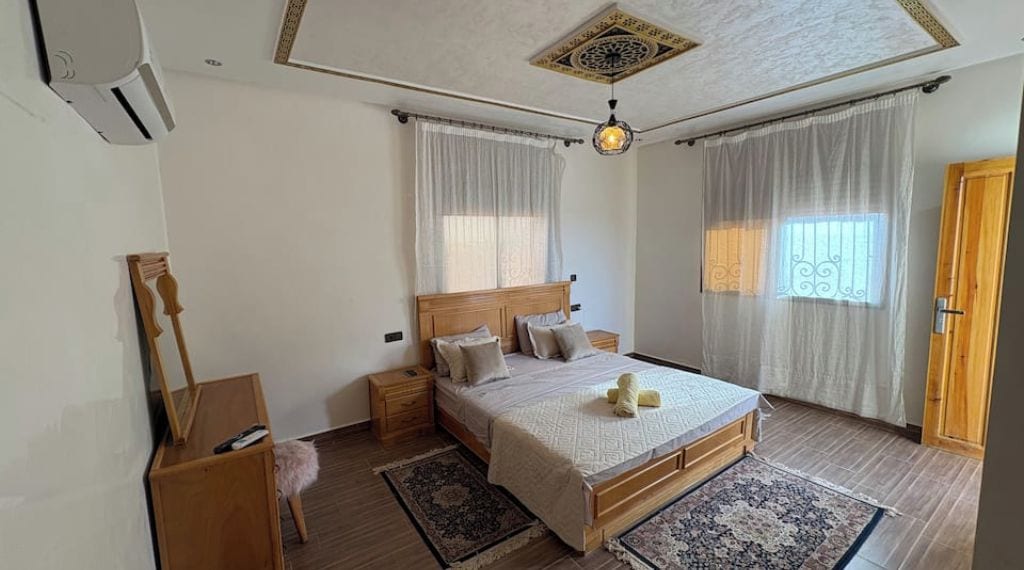 Una camera da letto con un letto di legno, un comodino, una lampada da tavolo, una sedia, una finestra con tende e un lampadario.
