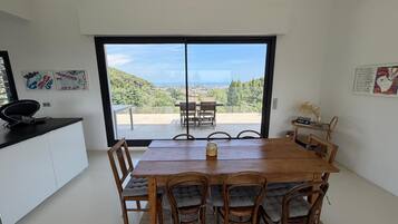 A dining area with a wooden table and chairs, a view of a hillside, and a kitchen area with a black countertop.
