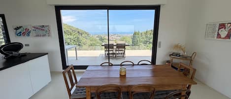 A dining area with a wooden table and chairs, a view of a hillside, and a kitchen area with a black countertop.