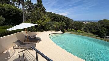 En swimmingpool med buet kant, omgivet af en belagt terrasse med liggestole og en parasol.
