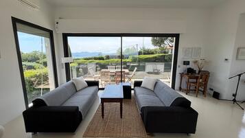A modern living room with two sofas, a coffee table, and a dining area with chairs and a table. Large windows provide a view of the outdoors.
