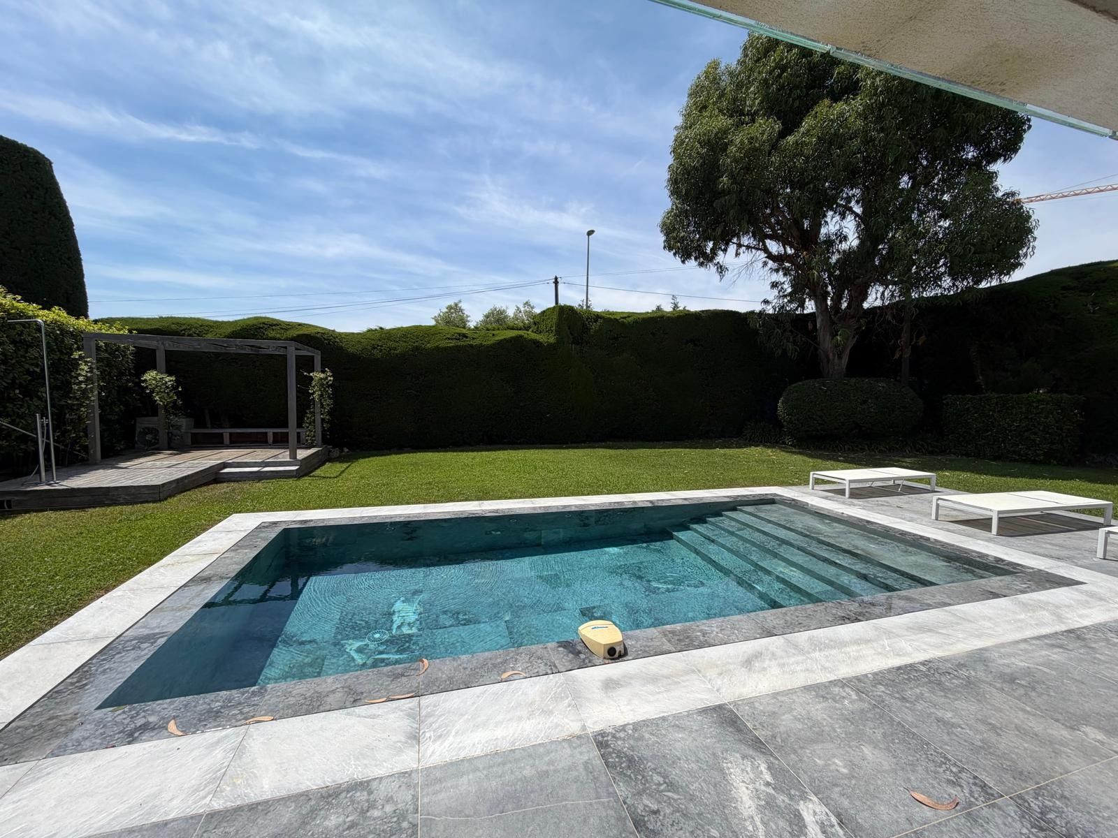 A rectangular swimming pool with a tiled deck, surrounded by a well-maintained lawn and a pergola structure.