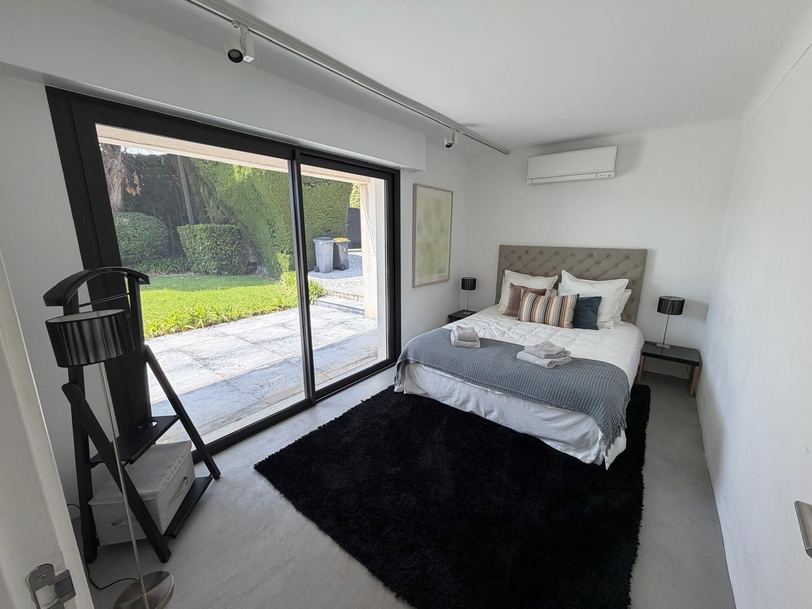 Una camera da letto moderna con un letto grande, un tappeto nero, una porta scorrevole in vetro che si apre su un giardino e una telecamera di sicurezza a soffitto.