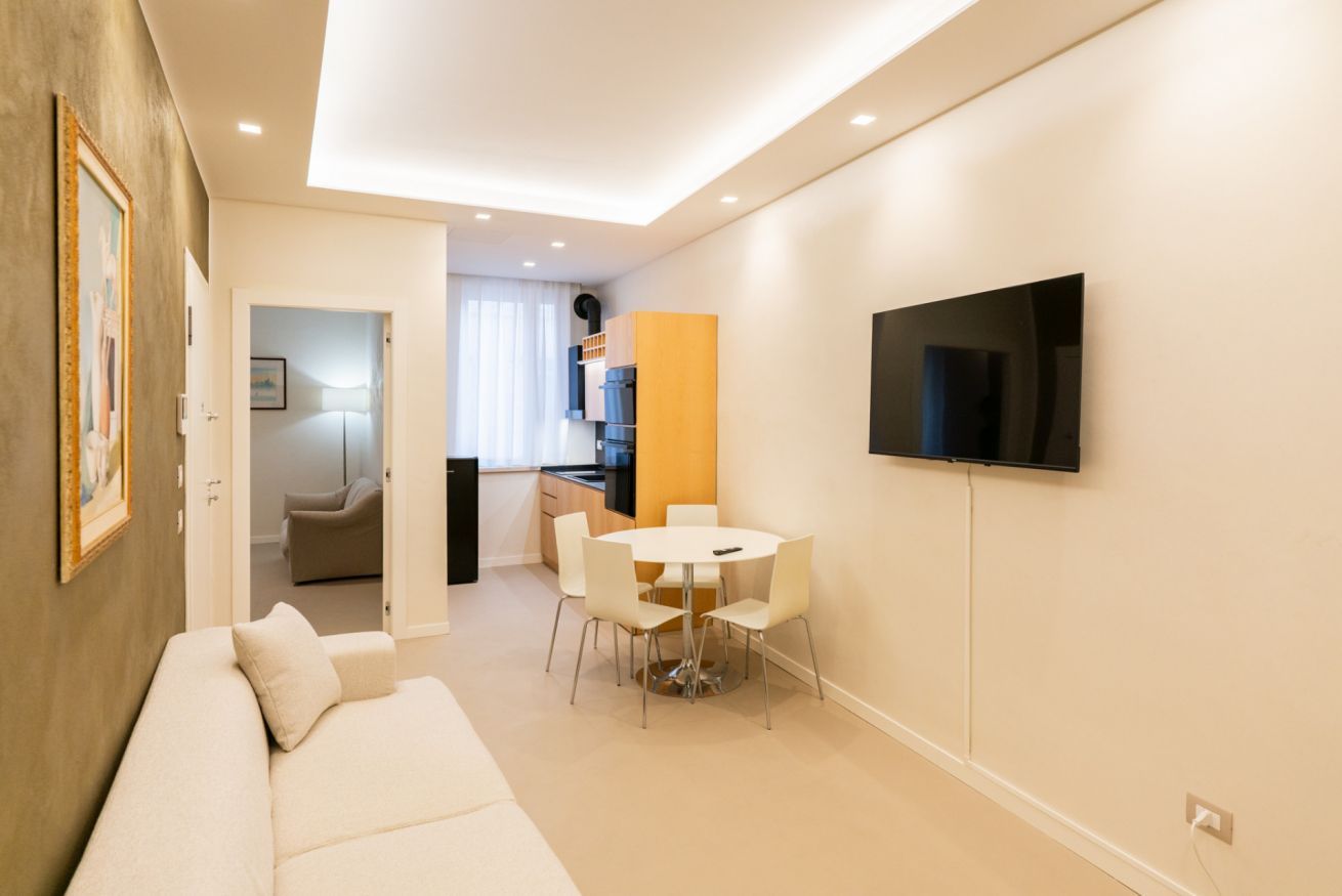 Un saló modern amb un sofà, una taula de dinar amb cadires, una zona de cuina i un televisió penjat.