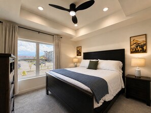 A bedroom with a bed, bedside lamps, a ceiling fan, and a window with a view of a parking lot and buildings. - Brand New Condo Steps to Lumberjack Square | Mt. LeConte View (Pigeon Forge)