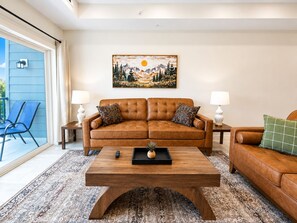 A living room with a brown leather sofa, two side tables with lamps, a coffee table, and a large painting on the wall. - Brand New Condo Steps to Lumberjack Square | Mt. LeConte View (Pigeon Forge)