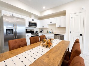 A modern kitchen with white cabinets, stainless steel appliances, and a wooden dining table with brown upholstered chairs. - Brand New Condo Steps to Lumberjack Square | Mt. LeConte View (Pigeon Forge)