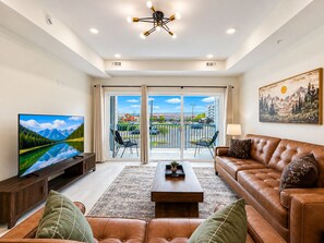 A modern living room with a television, leather sofa, and a balcony with a view of a parking lot and buildings. - Brand New Condo Steps to Lumberjack Square | Mt. LeConte View (Pigeon Forge)