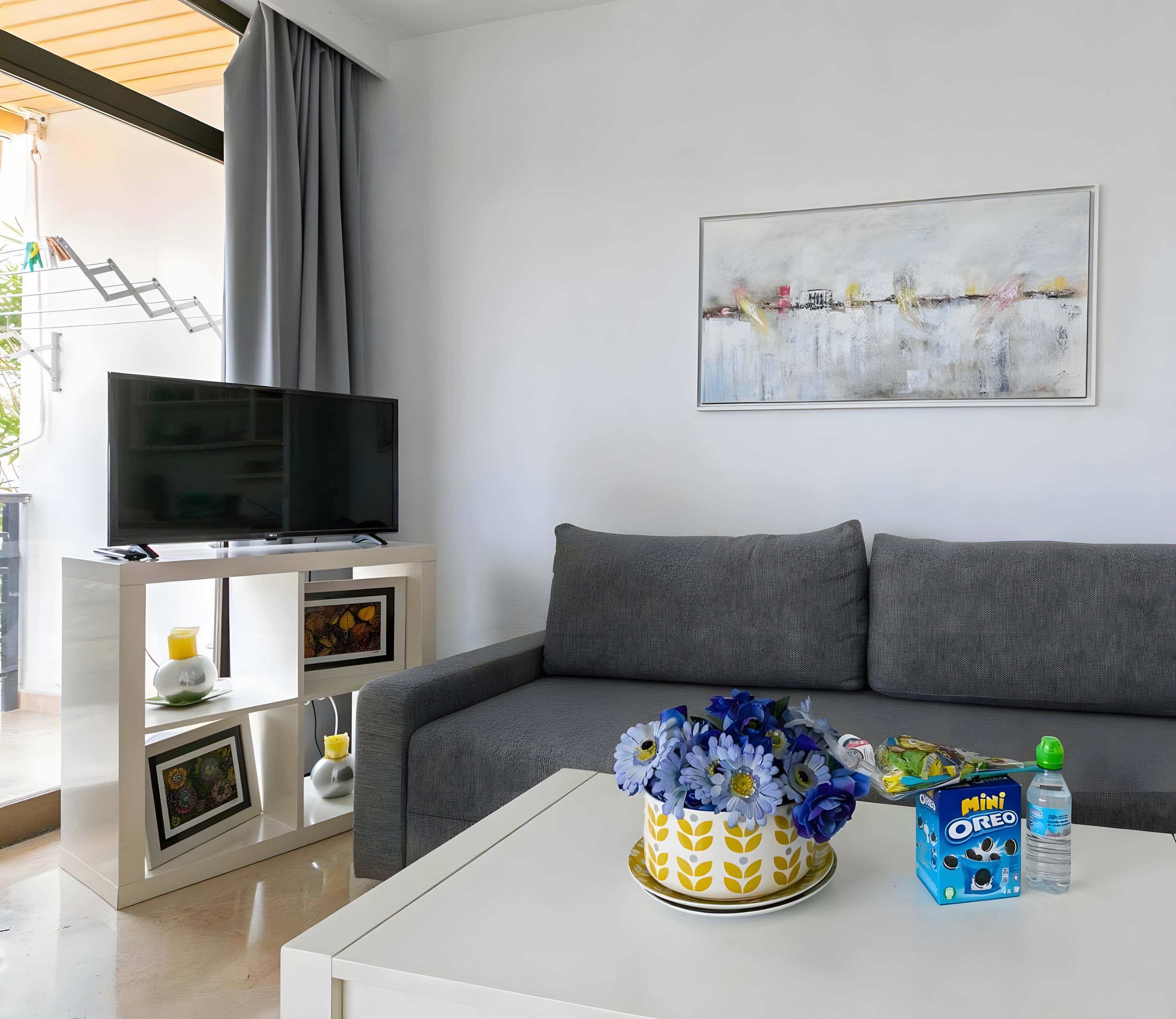 A modern living room with a grey sofa, a flat-screen TV, a white coffee table with a vase of flowers, and a painting on the wall.