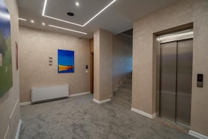 Egy folyosó, melynek a padlója szőnyeggel van bevonva, az egyik falán egy festmény található, és van benne egy lift.