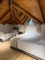 A bedroom with two beds, wooden ceiling beams, a window with curtains, and a bedside table with a lamp.