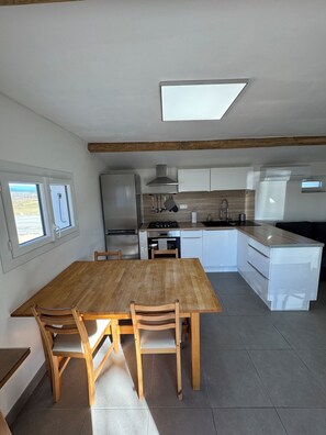 A modern kitchen with a dining area, featuring a wooden table and chairs, a built-in oven, and a large window. - Vacation Rental in the Heart of the Ardèche (Beaulieu)