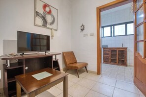 Una stanza con televisione su un supporto in legno, una sedia marrone, un tavolo di legno e vista su un balcone attraverso una porta aperta.