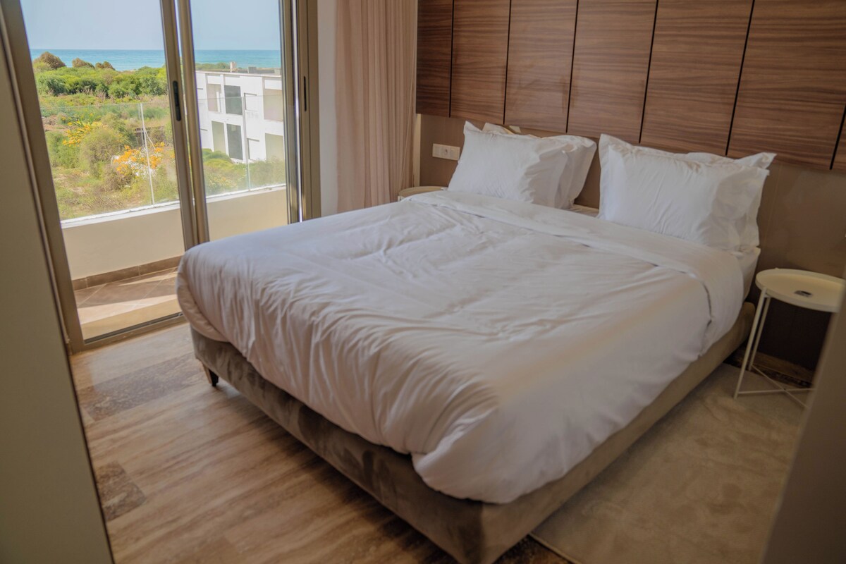 A bedroom with a large bed, a wooden headboard, and a view of the sea through a window.