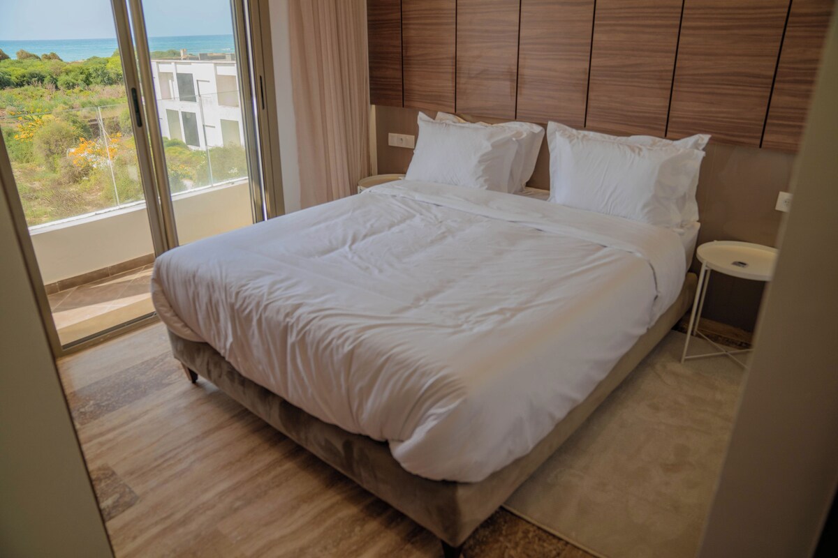 A hotel room with a large bed, a nightstand, and a view of the ocean.