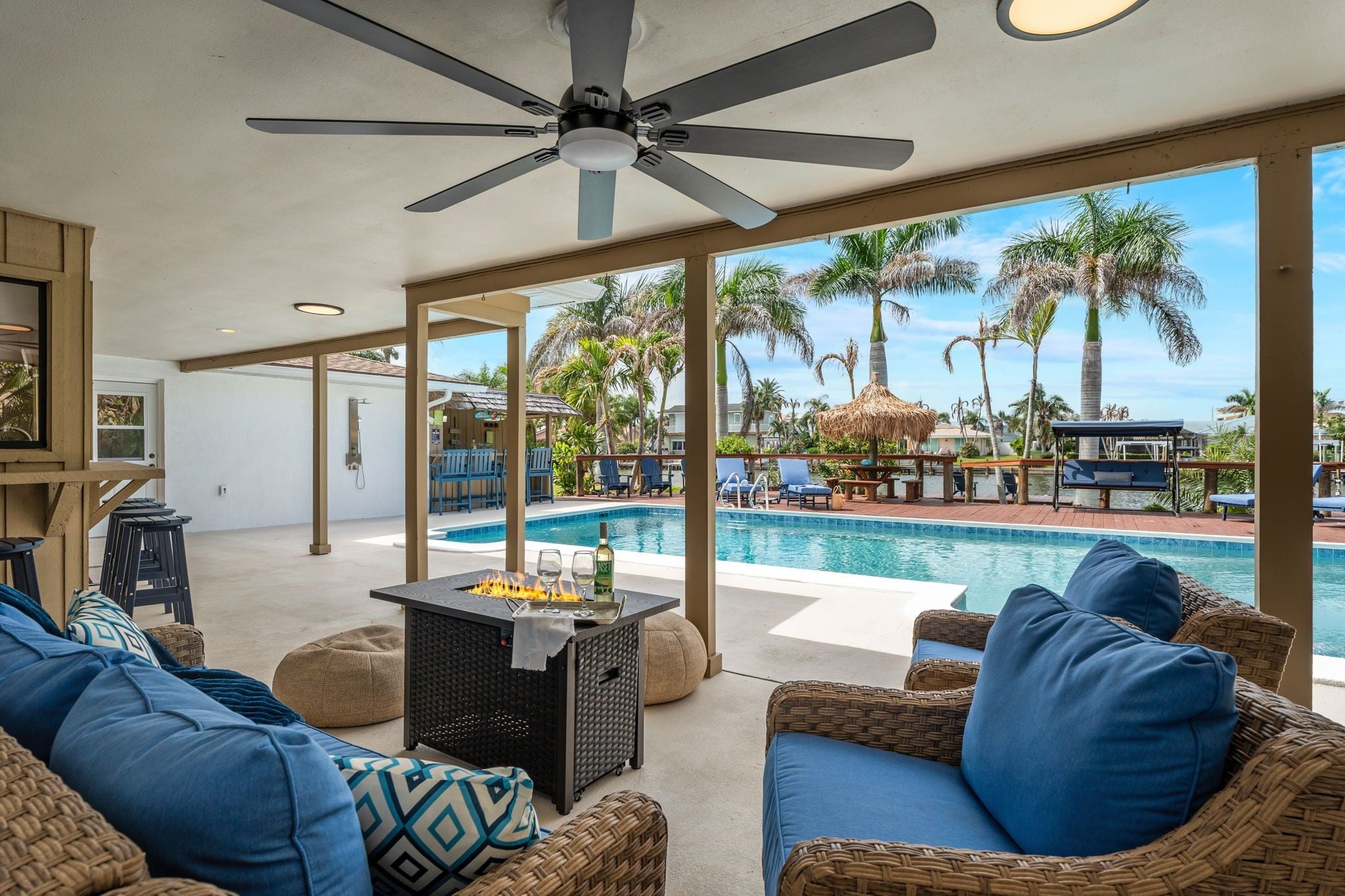 Un espace aménagé au bord de la piscine, avec des meubles en osier, un ventilateur de plafond et une vue sur la piscine et les palmiers.