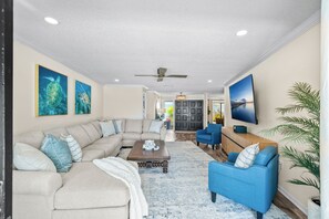 A spacious living room with a sectional sofa, a coffee table, and a blue armchair. There is a television mounted on the wall and a ceiling fan. - Waterfront 5BR Estate w/ Heated Pool | Walk to Cocoa Beach (Cocoa Beach)