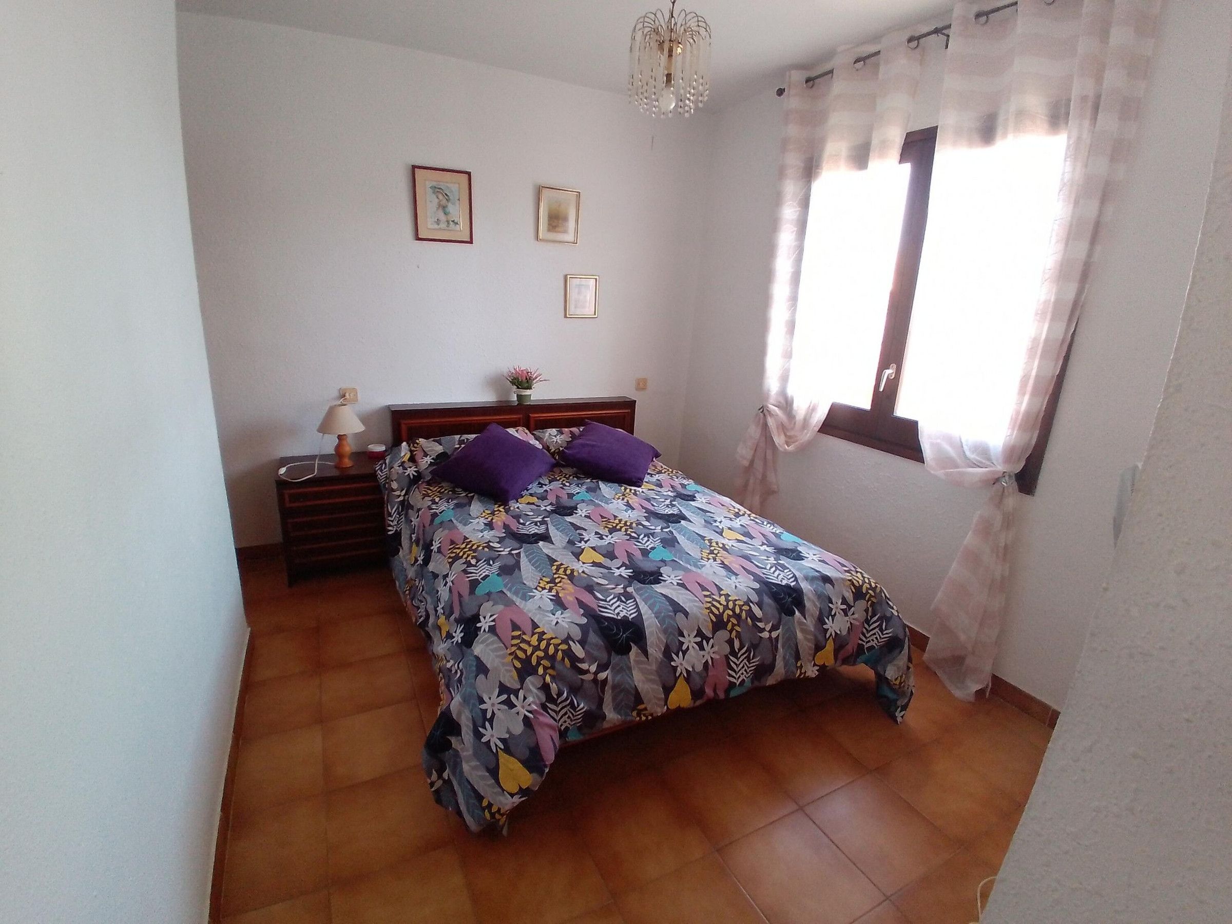 A bedroom with a bed, a nightstand, a lamp, a window with curtains, and a chandelier.