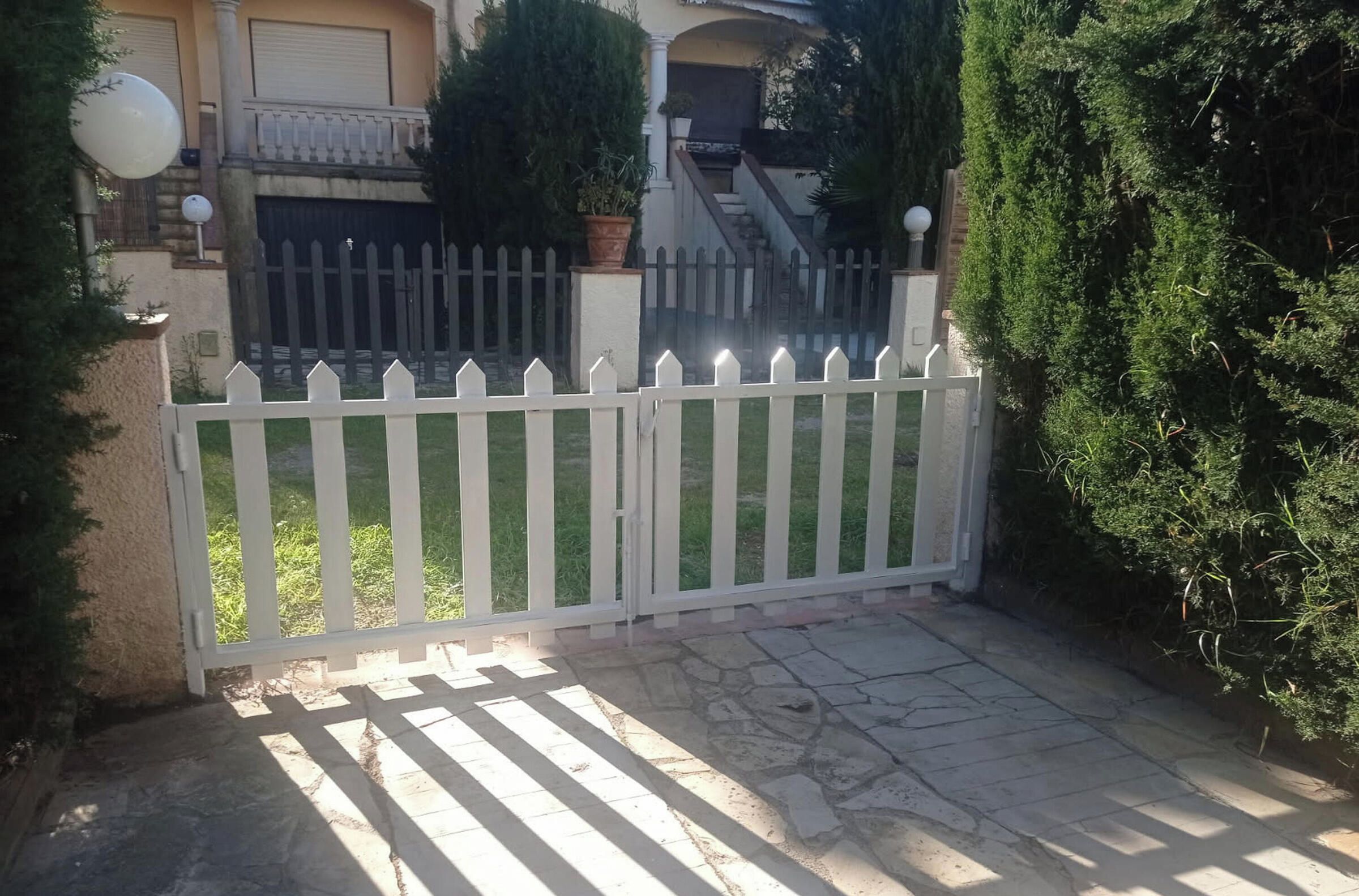 A gated entrance with a white picket fence, a paved driveway, and a house with a balcony and potted plants.
