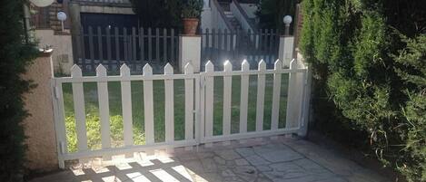 A gated entrance with a white picket fence, a paved driveway, and a house with a balcony and potted plants.