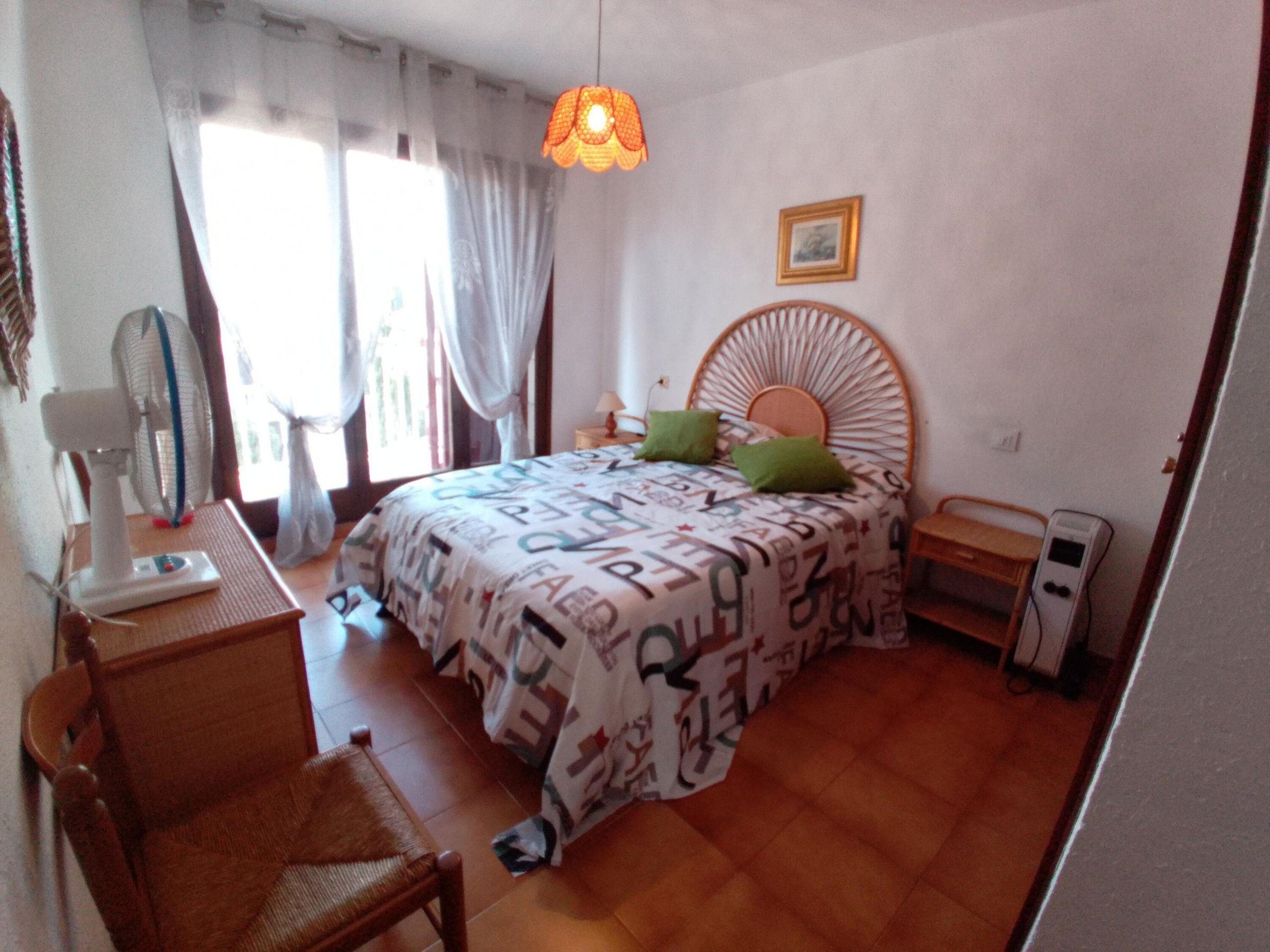 Ein Schlafzimmer mit Bett, Sessel, Nachttisch, Lampe, Ventilator und einem Bild an der Wand.