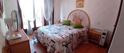 Un dormitorio con una cama, una silla, una mesita de noche, una lámpara, un ventilador y un cuadro en la pared.