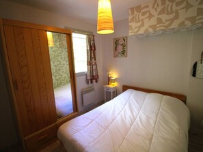A bedroom with a bed, a wooden wardrobe, a window with curtains, a bedside lamp, and a wall with a floral pattern. - Quiet apartment with parking, pets OK - 4 guests, mountain view (Bagnères-de-Luchon)