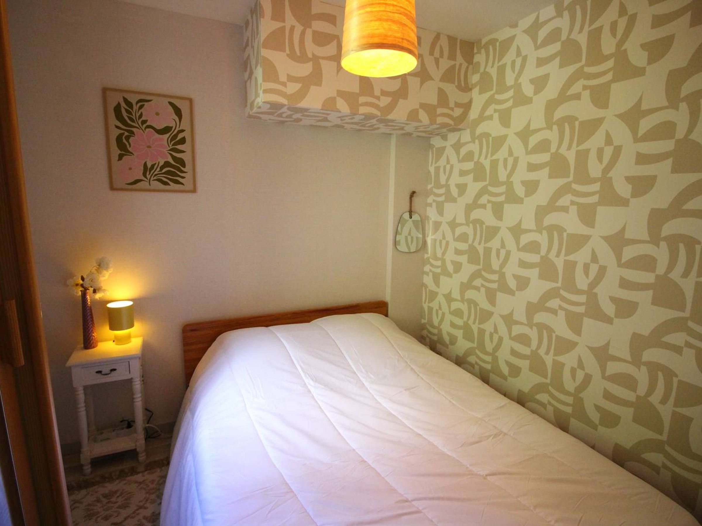 A bedroom with a bed, a bedside lamp, a wall-mounted mirror, and a floral-patterned wall.