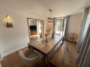 Een eetgedeelte met een houten tafel, een bank, een stoel, een vaas met gedroogde bloemen en een wandlamp.
