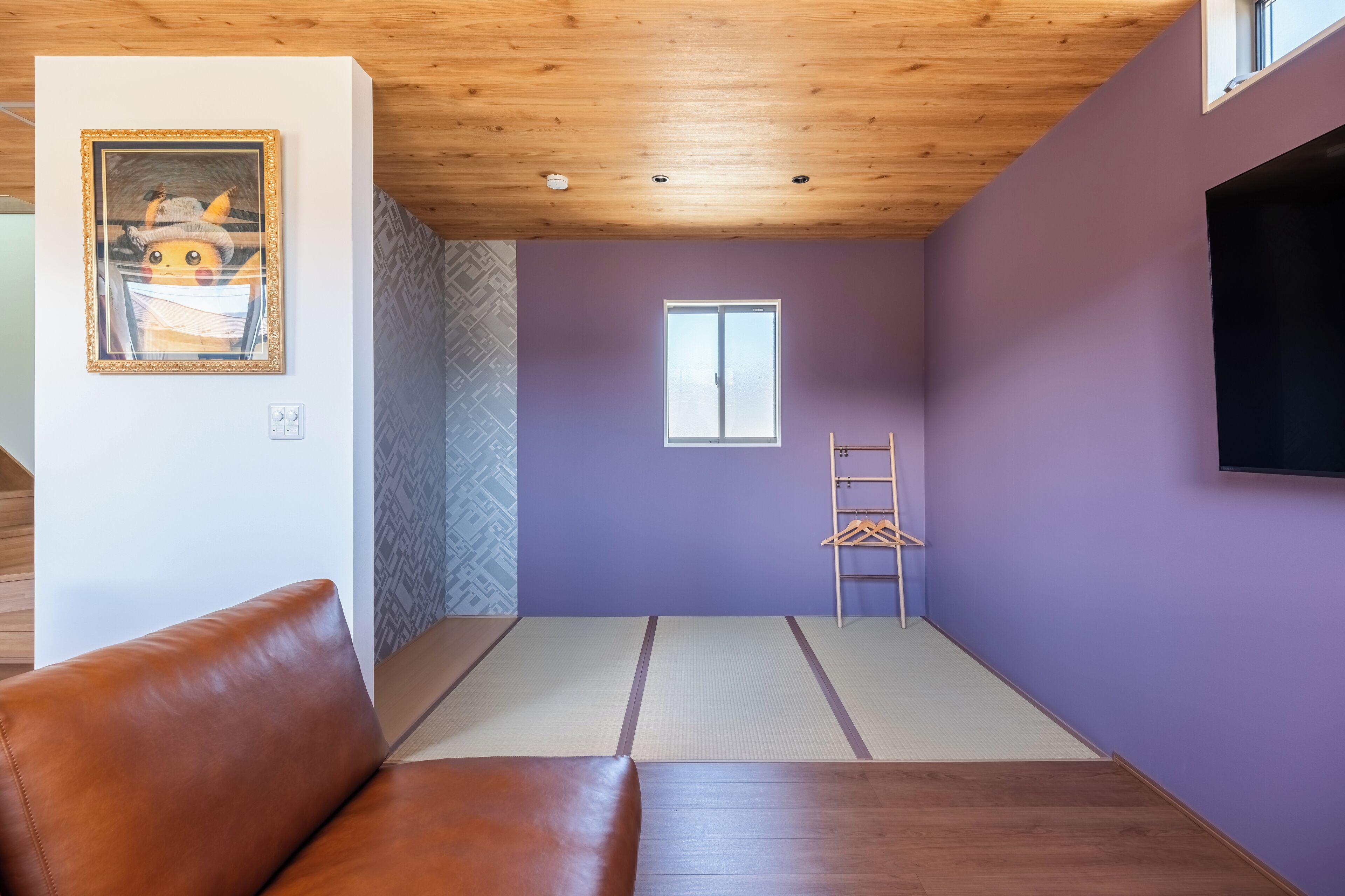 A room with a brown leather sofa, a wooden ceiling, a purple wall, a window, and a TV mounted on the wall.