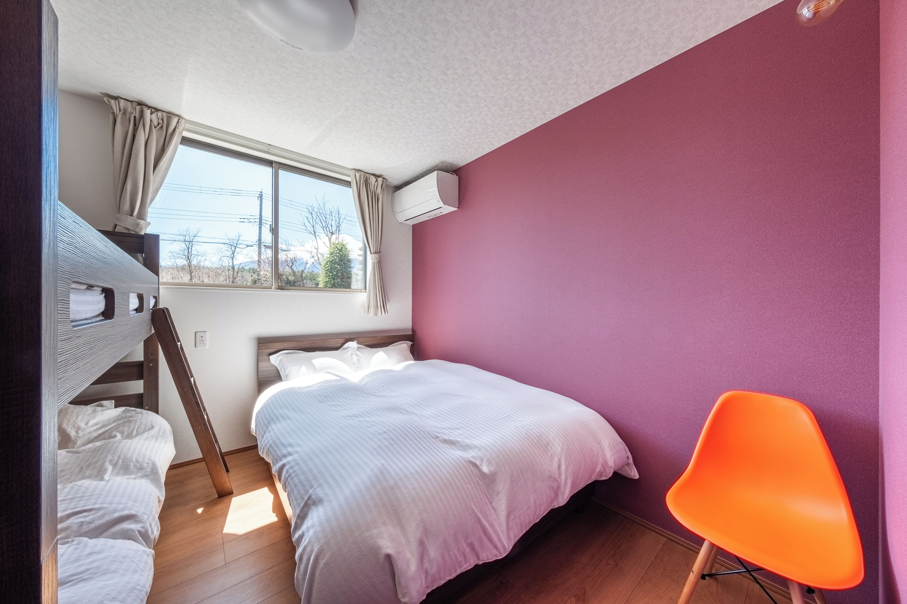 Ein Schlafzimmer mit Etagenbett, einem Einzelbett, einem Fenster mit Vorhängen, einer Klimaanlage und einem leuchtend orangefarbenen Sessel.