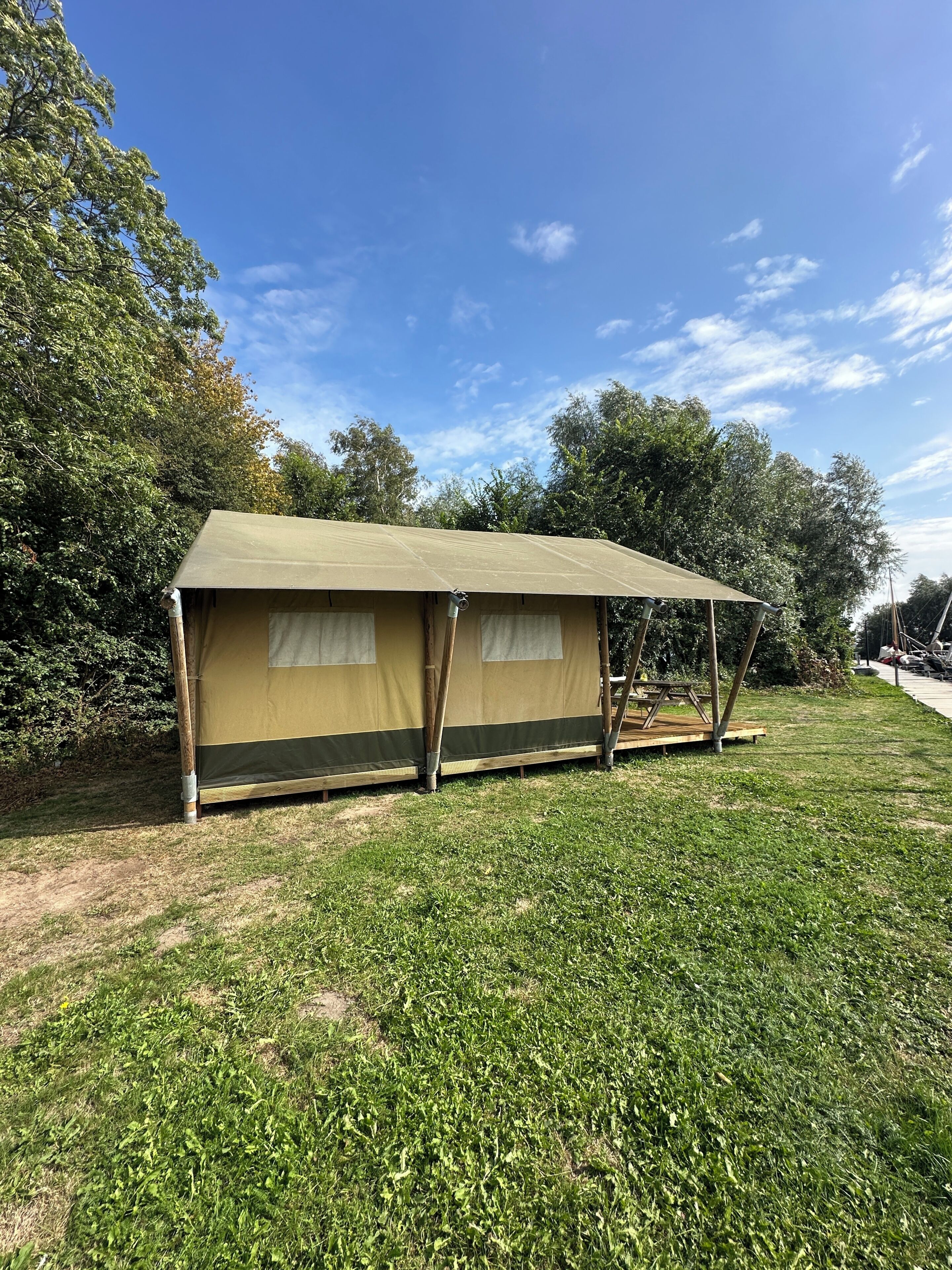 Una tienda de una sola habitación con techo de lona y estructura de madera, ubicada en un campo cubierto de hierba con árboles al fondo.