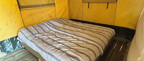 Un lit simple avec un matelas à rayures, dans une pièce qui ressemble à une tente, aux murs jaunes et au plancher en bois.
